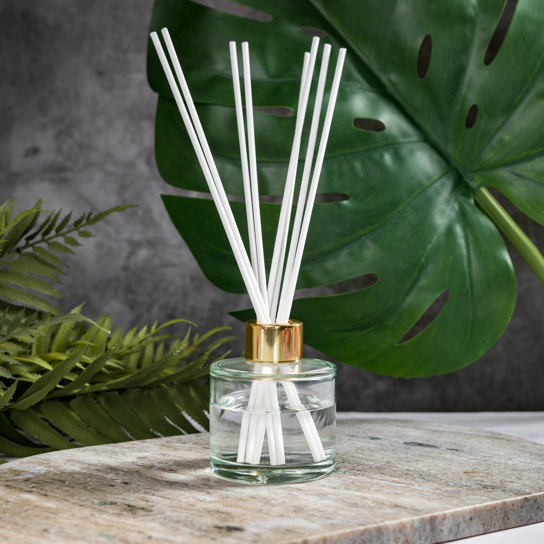 Reed diffuser with white sticks in a clear bottle with gold cap on a wooden surface with green leaves in the background