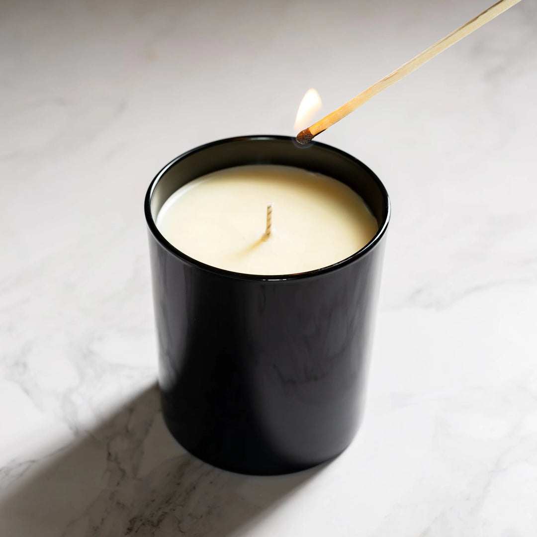Black candle in a glass container with a lit wick on a marble surface